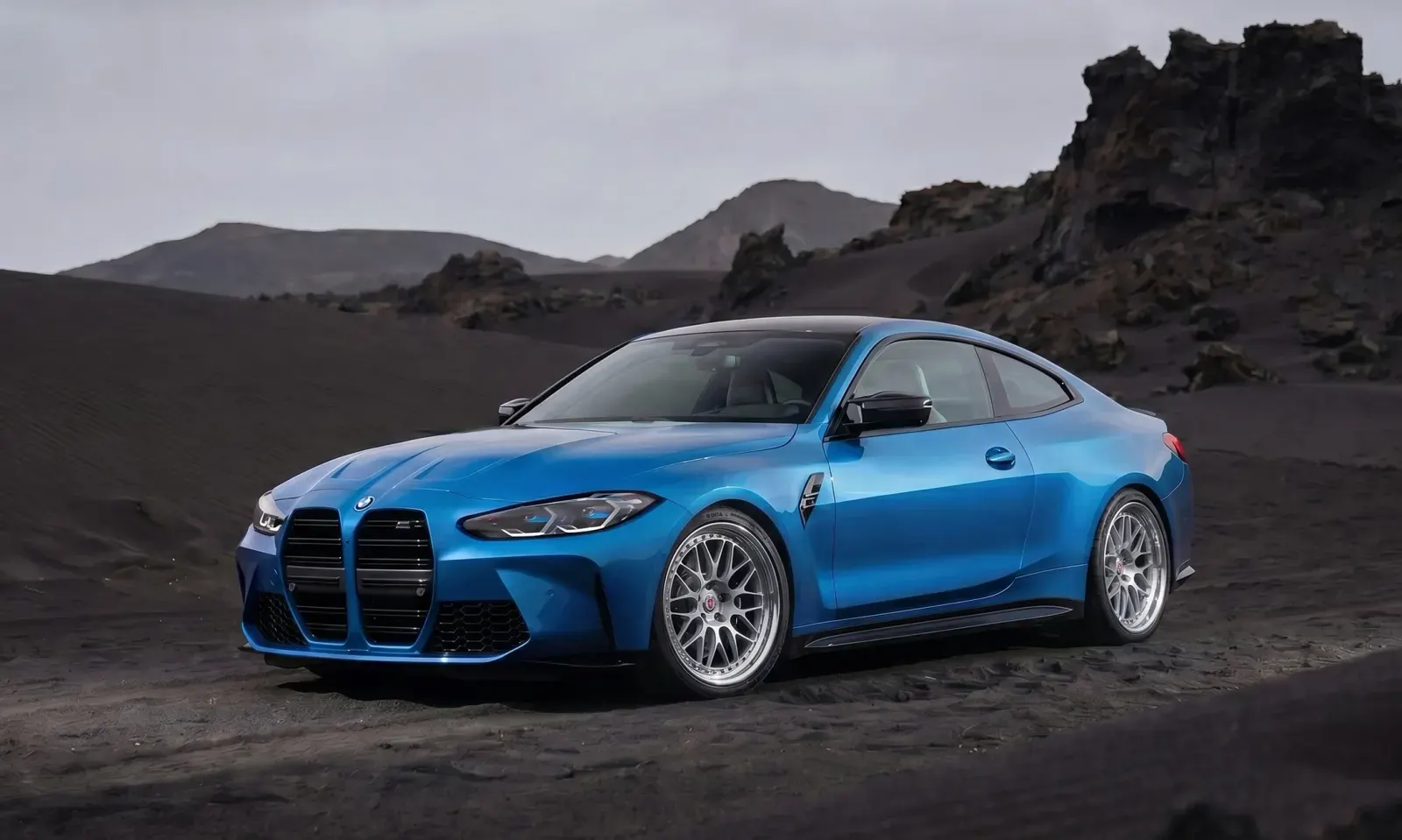 Blue BMW M4 coupe with bright multi-spoke performance wheels on black volcanic sand