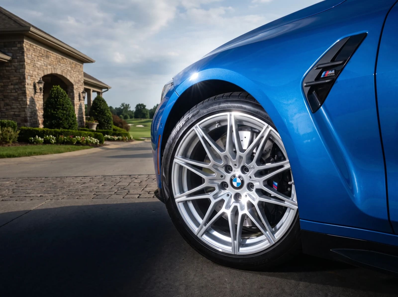 Blue BMW M4 close-up with silver factory-style multi-spoke wheels outside a stone home